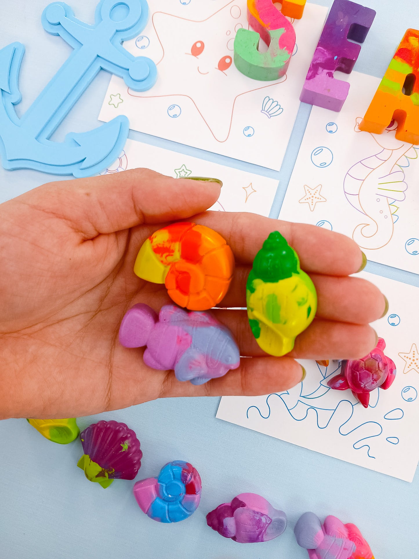 Hand holding multicolor sea creature crayons shaped like shells and fish, with ocean-themed coloring cards and SEA letter crayons in background; handmade non-toxic crayons for kids’ gifts and party favors.
