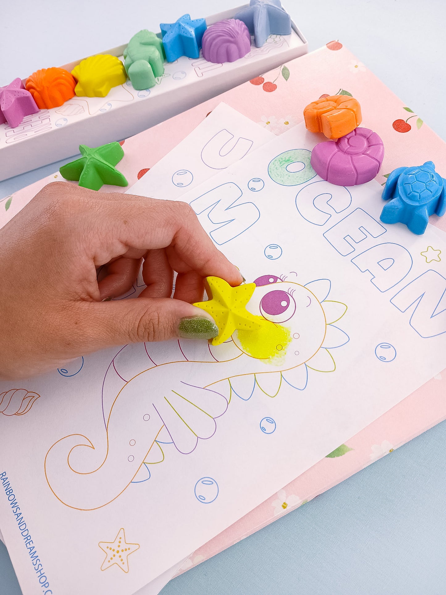 Hand holding yellow starfish crayon with other sea shapes displayed on pastel ocean coloring card, handmade in USA.